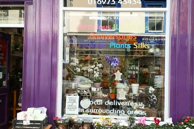 storefront.jpg Exterior of a florist shop on King Street, Wigton. The building front is painted a bright purple. Text on the large window advertises services. The phone number is also displayed. The window is full of green plants and flowers in pots. Outside, on the sidewalk, are trays of potted flowers and a small table holding a crate of greeting cards.